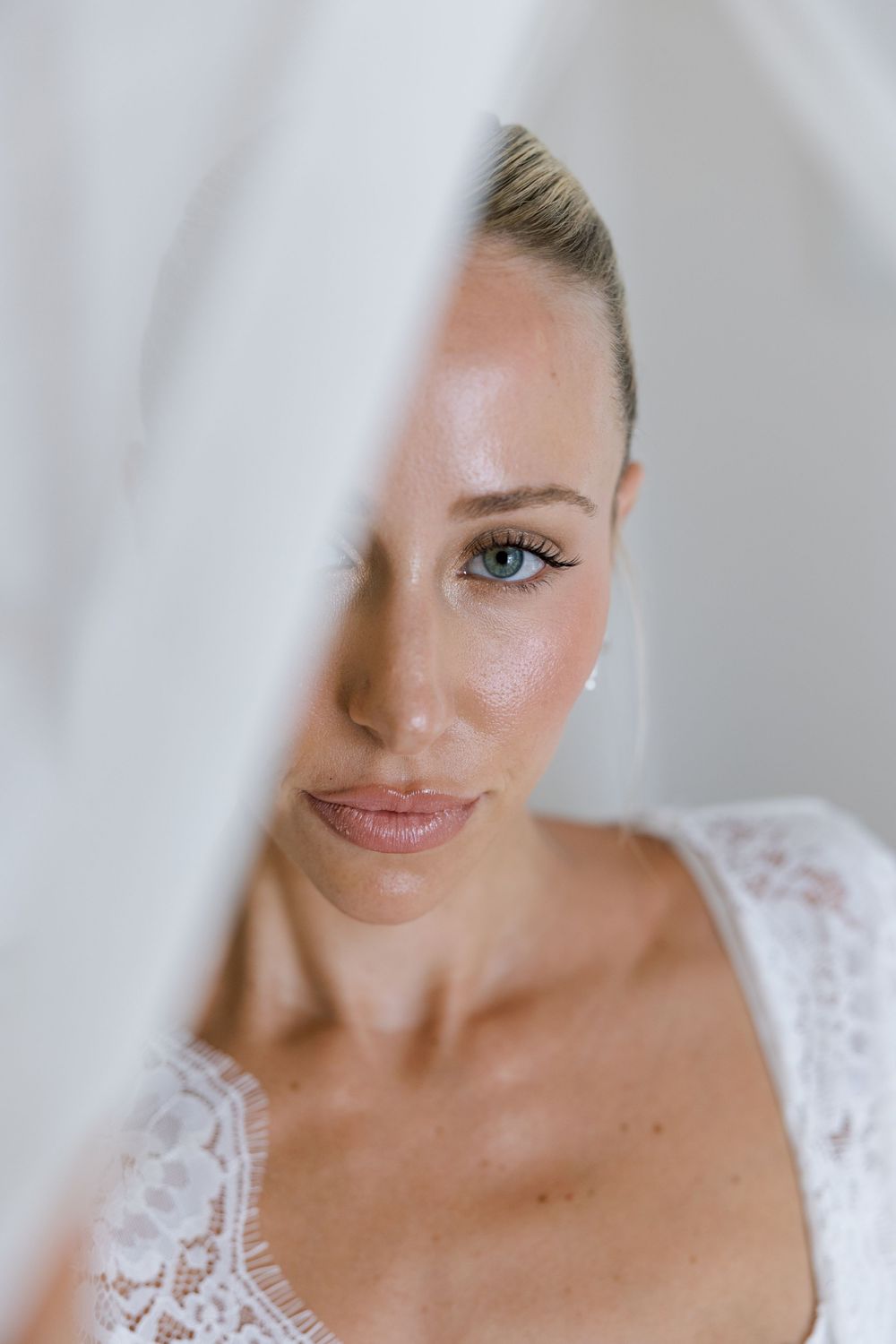 Close-up portrait of a blonde bride with blue eyes in white lace dress, partially obscured by sheer veil.