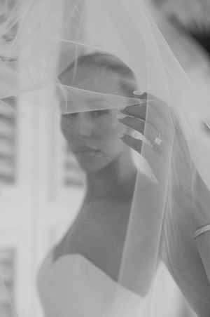 Black and white portrait of a bride partially veiled, holding her wedding veil with a serene expression.