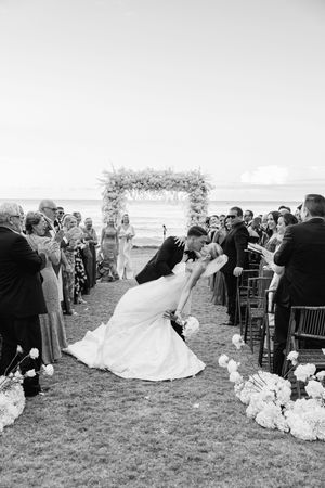 Black and white photo of bride and groom sharing a romantic dip kiss at outdoor beach wedding ceremony.