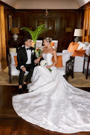 Bride and groom sit together in elegant wood-paneled room, her lace gown trailing gracefully, surrounded by orange accent pillows.