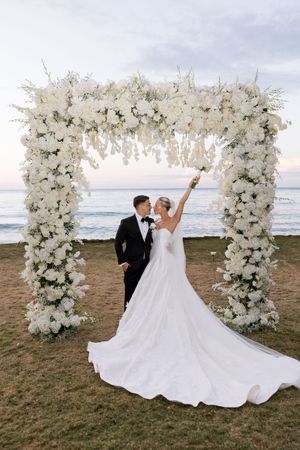 Bride and groom pose under a white floral arch by the ocean at sunset, bride raising her bouquet joyfully.