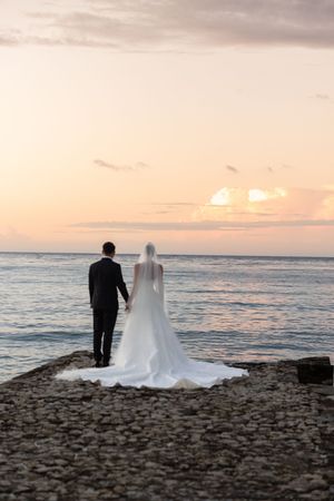 Bride and groom holding hands on rocky shoreline at sunset, overlooking calm ocean waters.