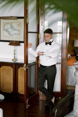 Groom in tuxedo adjusting cufflinks before wedding ceremony in elegant room with wooden decor.