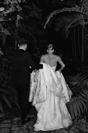 Black and white photo of bride and groom walking through lush tropical garden, bride in elegant backless lace gown.