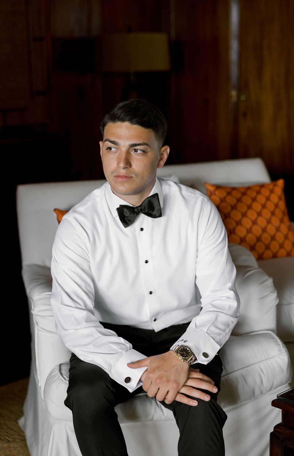 Young man in white tuxedo shirt and black bow tie sits confidently in white armchair wearing a gold watch.