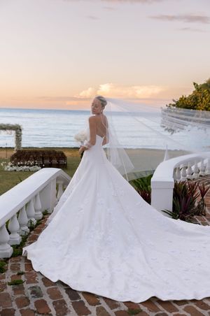 Bride in elegant white gown with long train and veil poses on terrace at sunset overlooking the ocean.