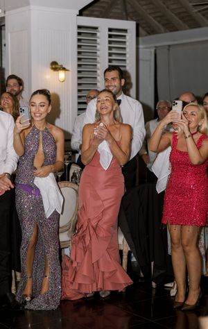 Elegantly dressed wedding guests in formal attire applaud and capture moments on phones at an upscale reception.