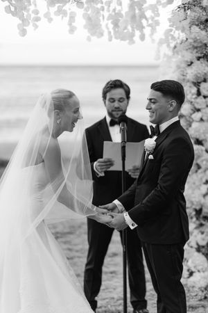 Black and white photo of a smiling bride and groom exchanging vows at an outdoor wedding ceremony by the ocean.