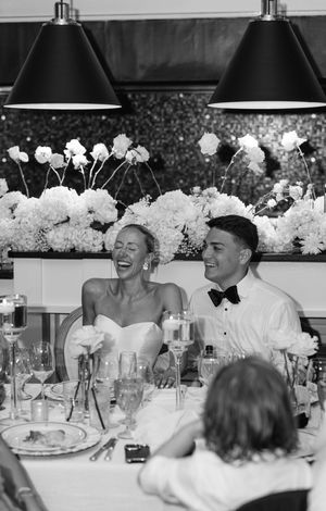 Black and white photo of a laughing bride and groom seated at their wedding reception table with floral arrangements.