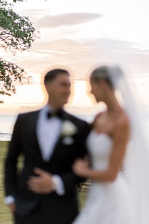 Blurred wedding couple in elegant attire share a romantic moment outdoors at sunset near a waterfront.
