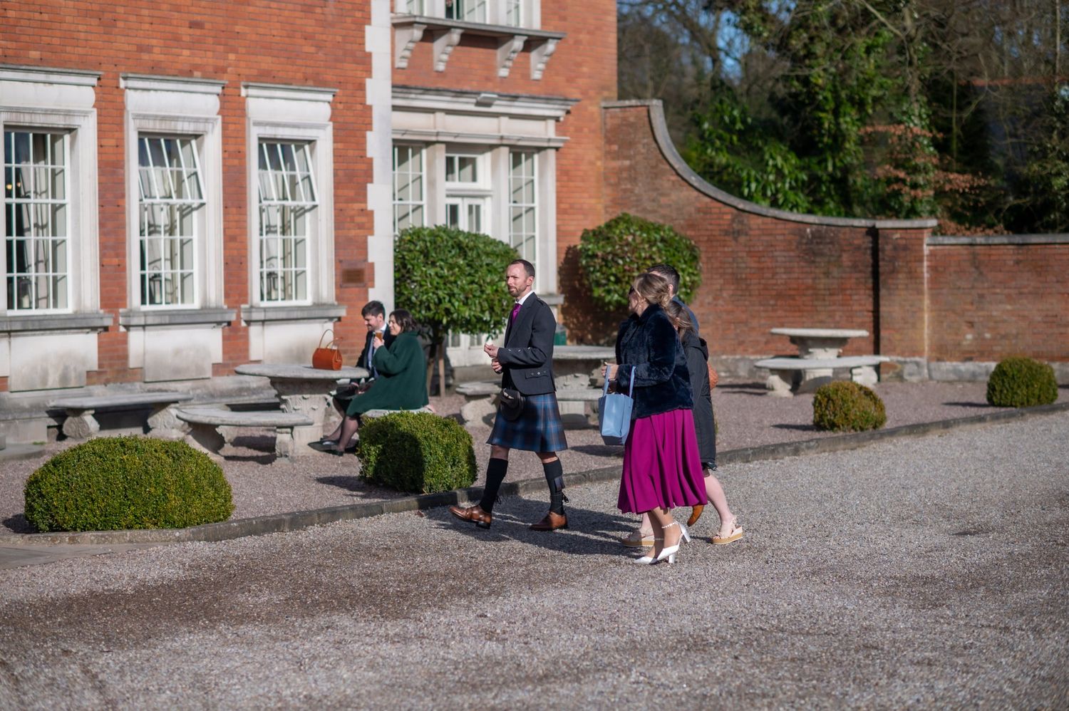 Elegantly dressed group walking outdoors near a brick building with manicured hedges on a sunny day.