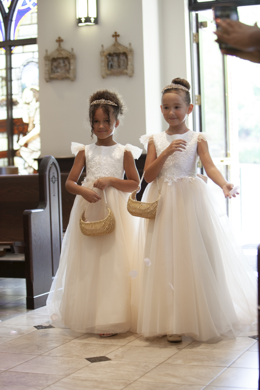 Two flower girls in white tulle dresses with wicker baskets pose in a church aisle during a wedding ceremony.
