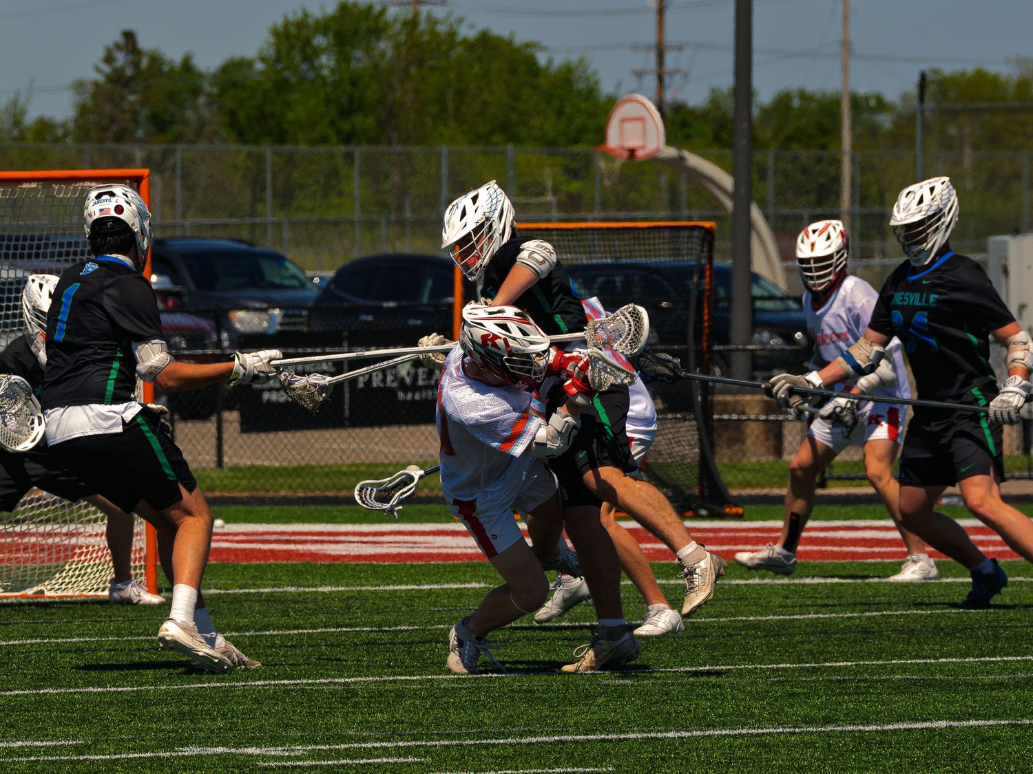 Janesville lacrosse player in black defends as a red and white opponent drives past with the ball on a sunlit field.