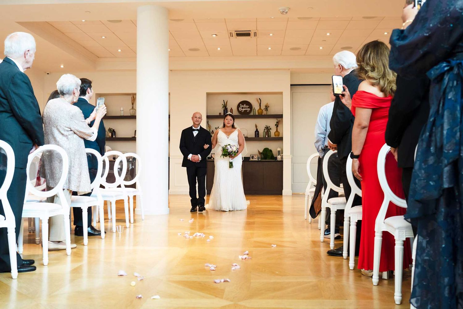 Bride walks down the aisle with escort at an elegant indoor wedding ceremony with white chairs.