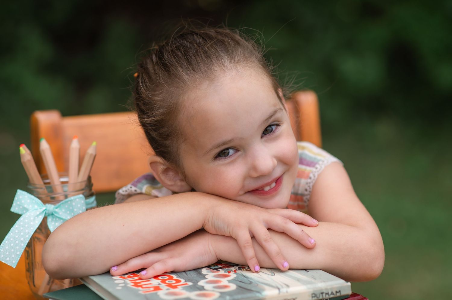 Smiling young girl resting her chin on her arms on a table, holding a book outdoors.