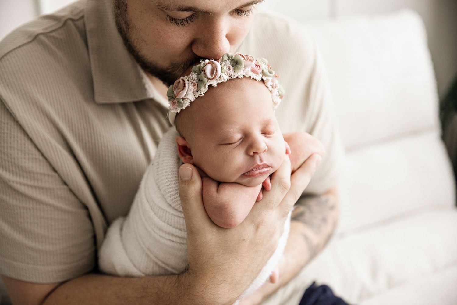 Father tenderly kisses sleeping newborn baby girl wearing floral headband wrapped in white swaddle.