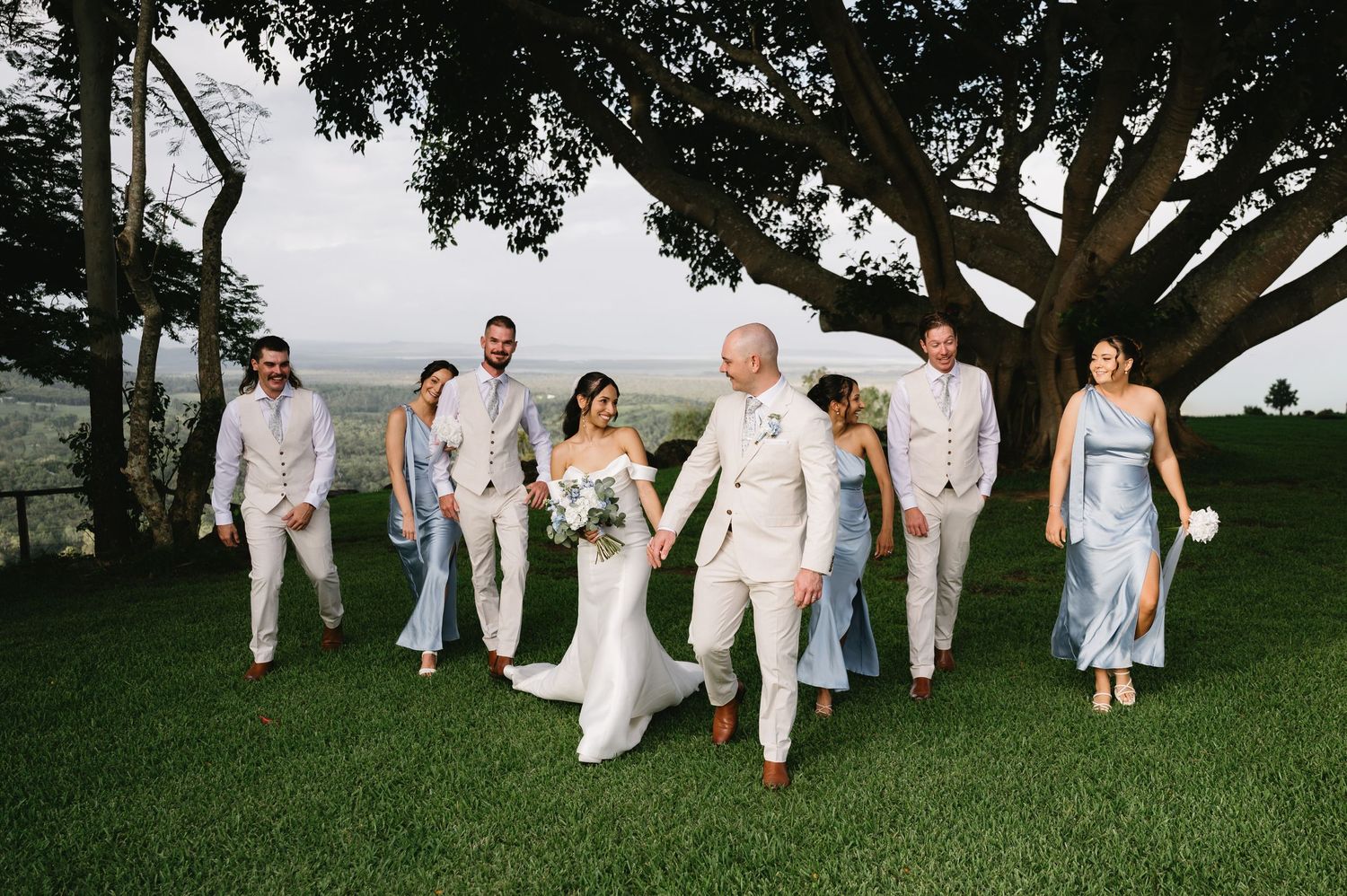 Wedding party walks on lush green lawn under large tree, bride in white gown with groom and bridal party in blue and beige.