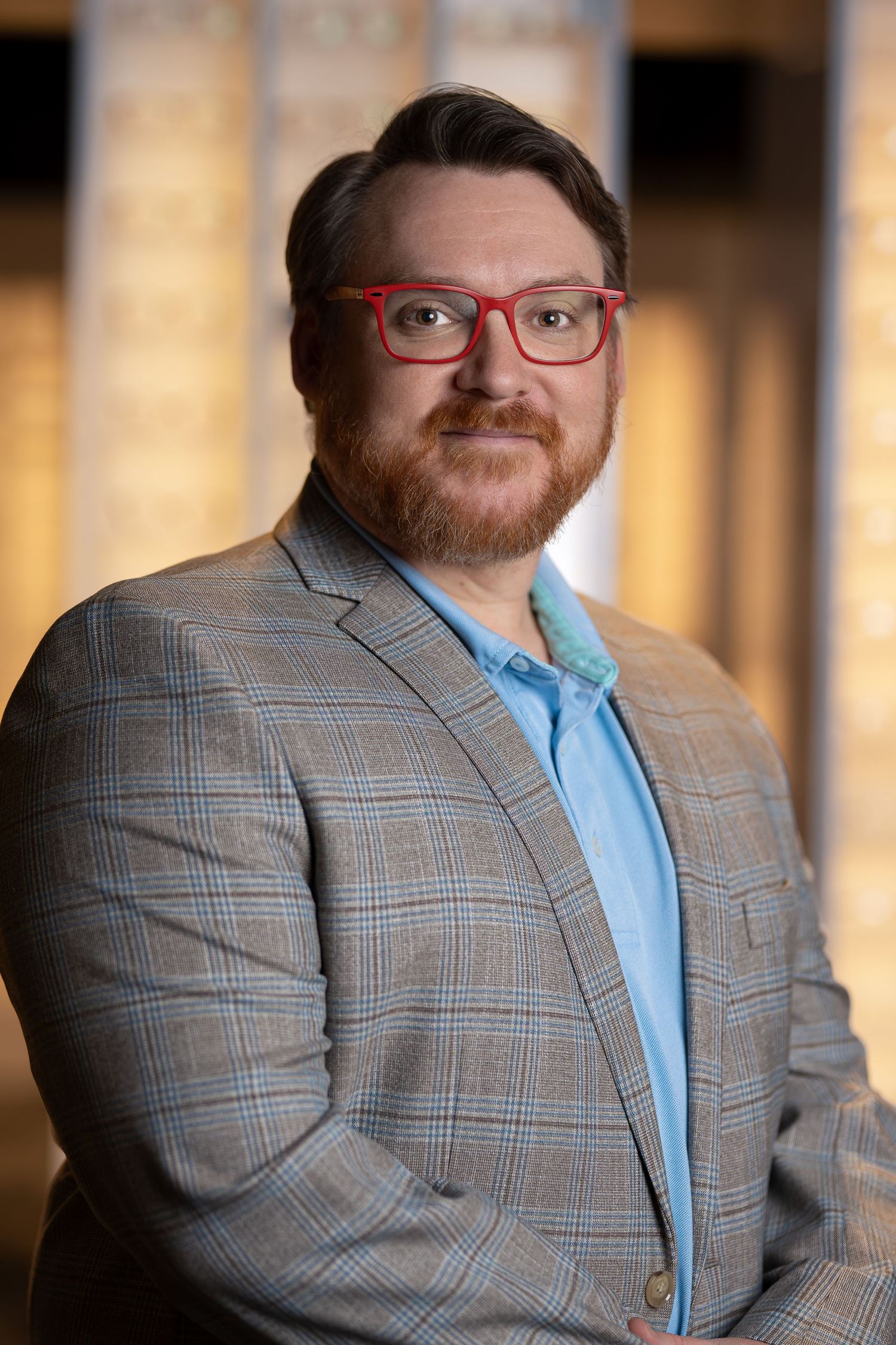 Professional man with red glasses and beard wearing a plaid blazer posing for corporate headshot.
