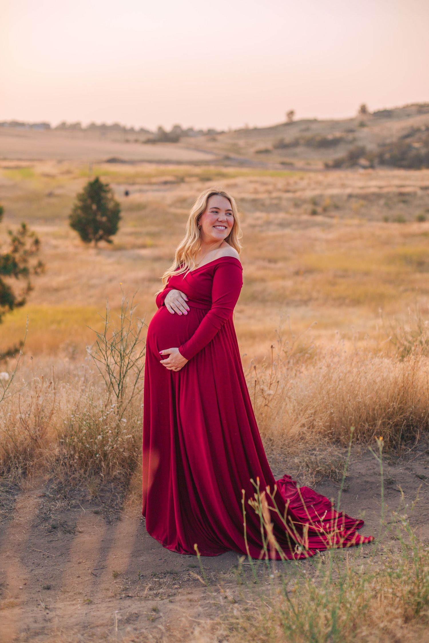 Smiling pregnant woman in elegant red maternity gown posing outdoors in golden field at sunset.