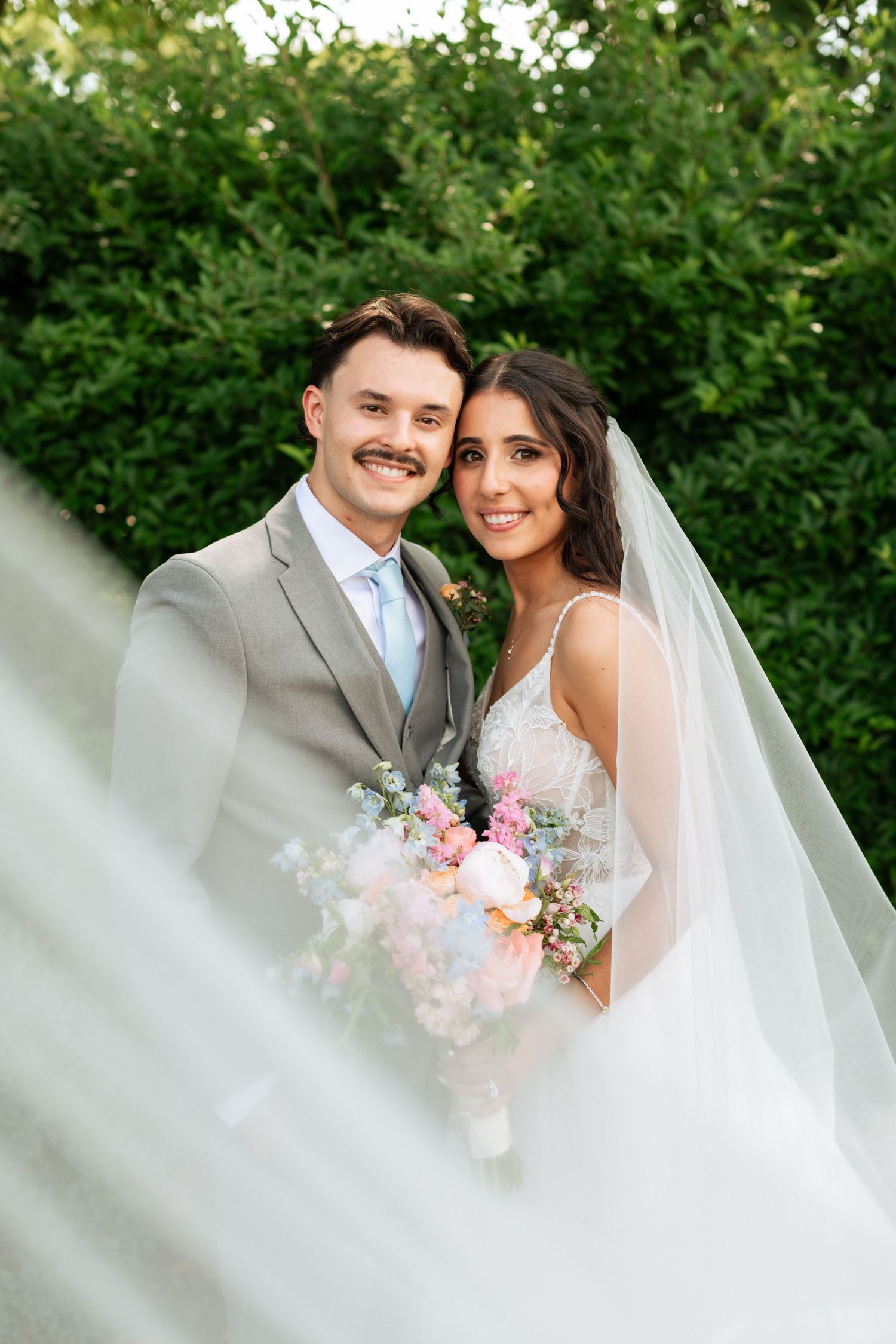 Bride and groom smiling outdoors, holding a pastel bouquet, with a flowing veil and lush green hedges in the background.