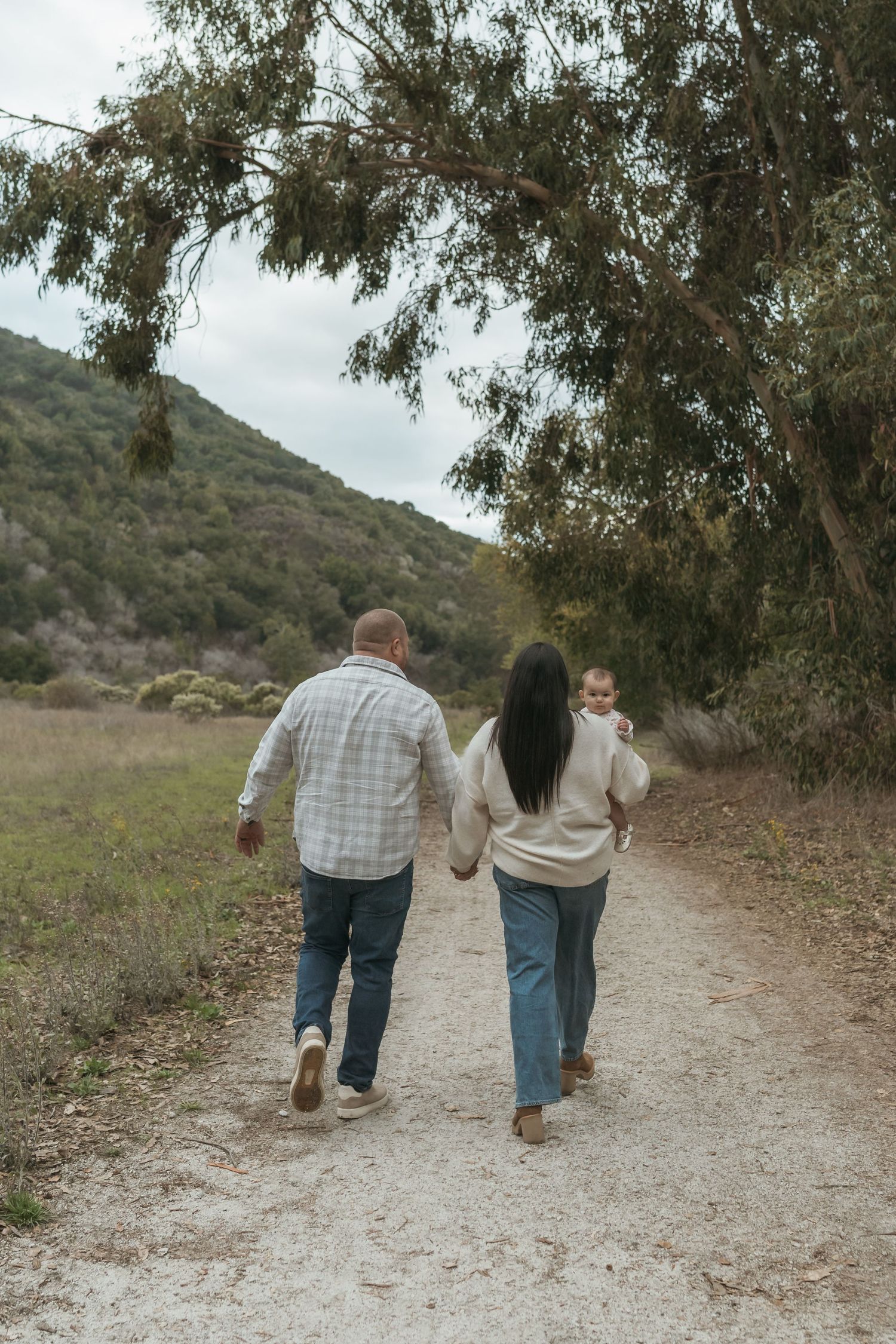 A family of three walks hand-in-hand down a scenic dirt path surrounded by trees and green hills.