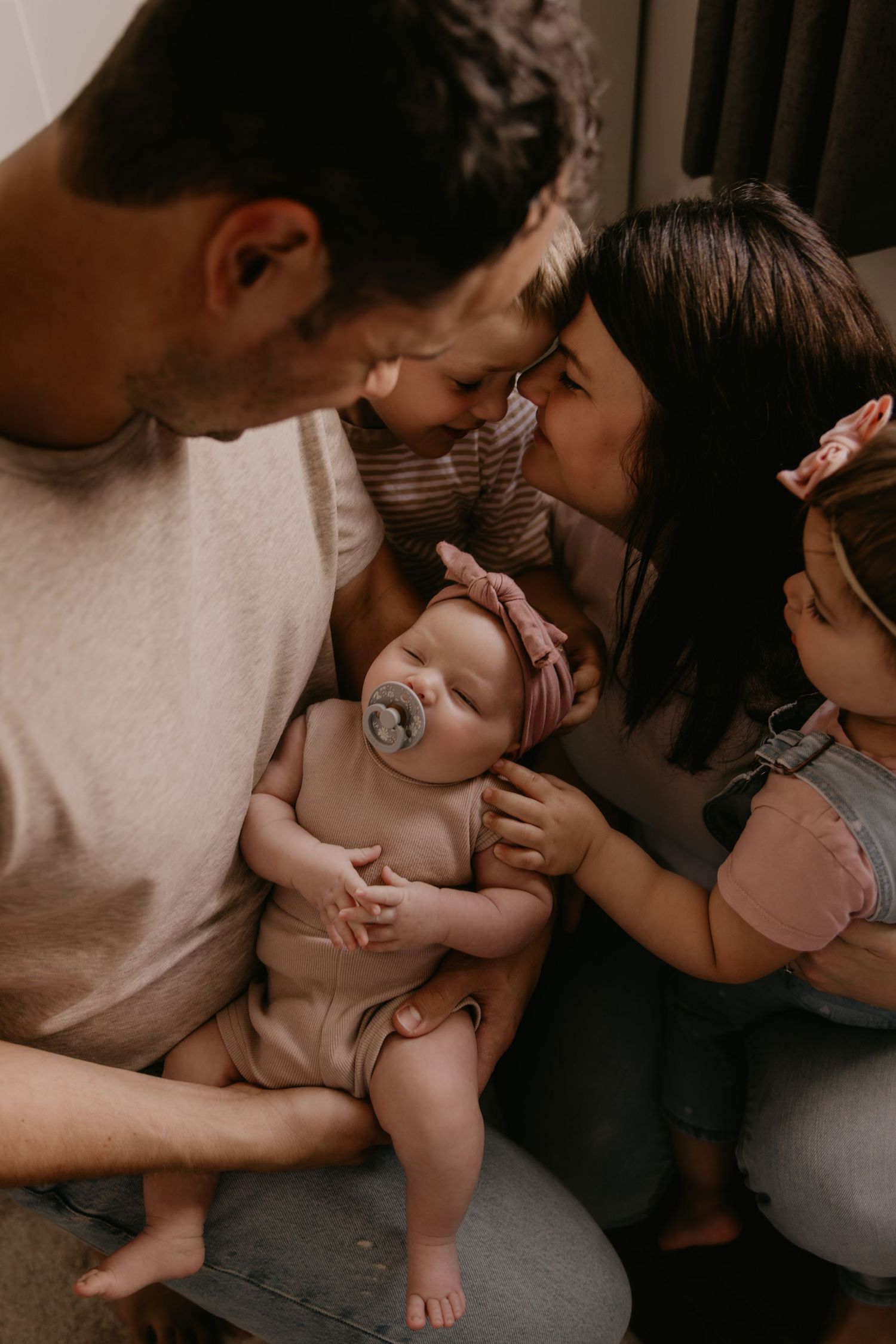 A loving family of four admires their newborn baby with a pacifier and pink headband in a warm, intimate moment.