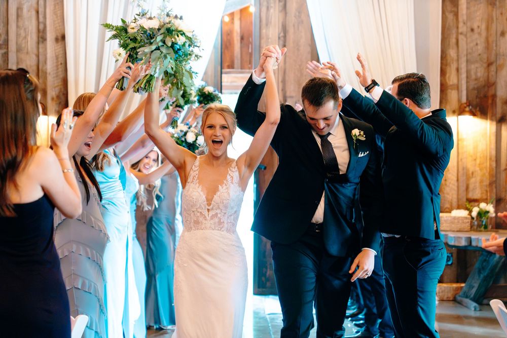 Bride and groom celebrate joyfully with wedding party cheering in elegant barn venue.
