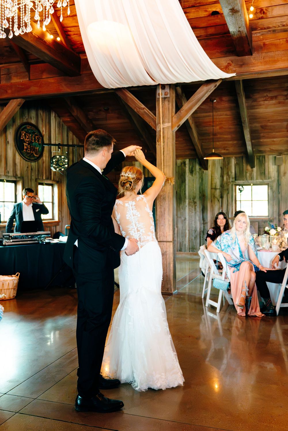 Bride and groom share first dance in rustic barn wedding venue with wood beams, string lights, and white draping.