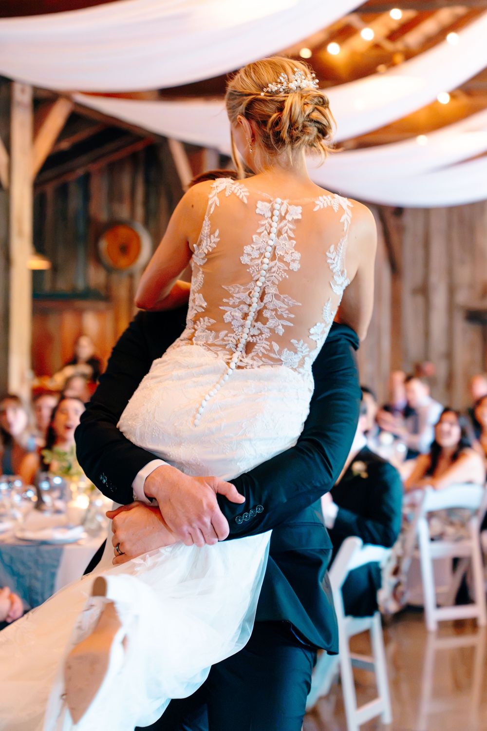 Bride and groom share first dance in rustic barn venue, showcasing stunning lace illusion back wedding gown.