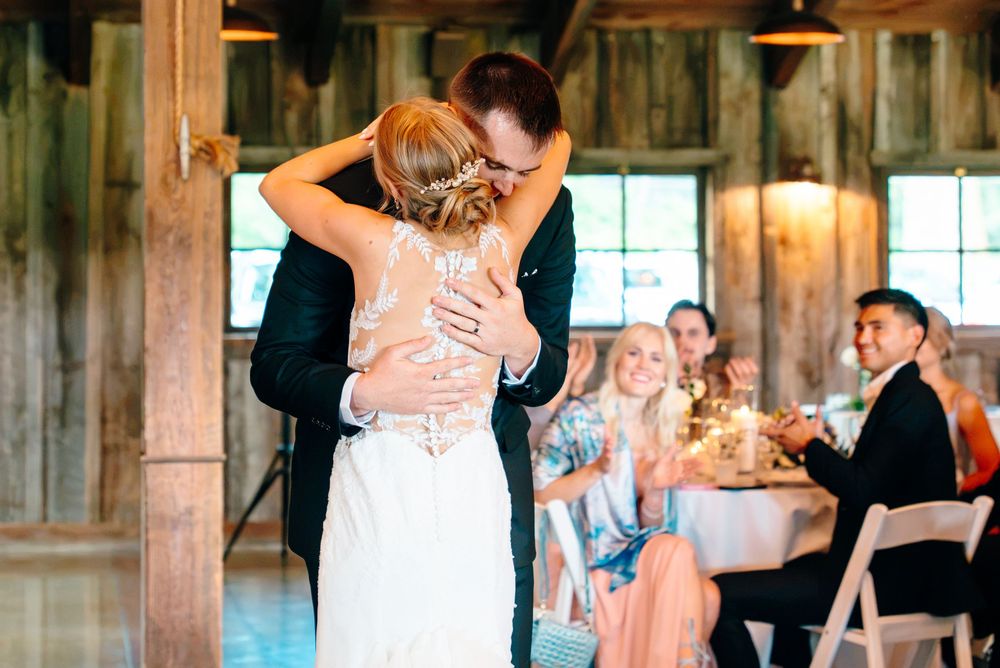 Bride and groom share first dance embrace in rustic barn wedding venue with guests watching in background.