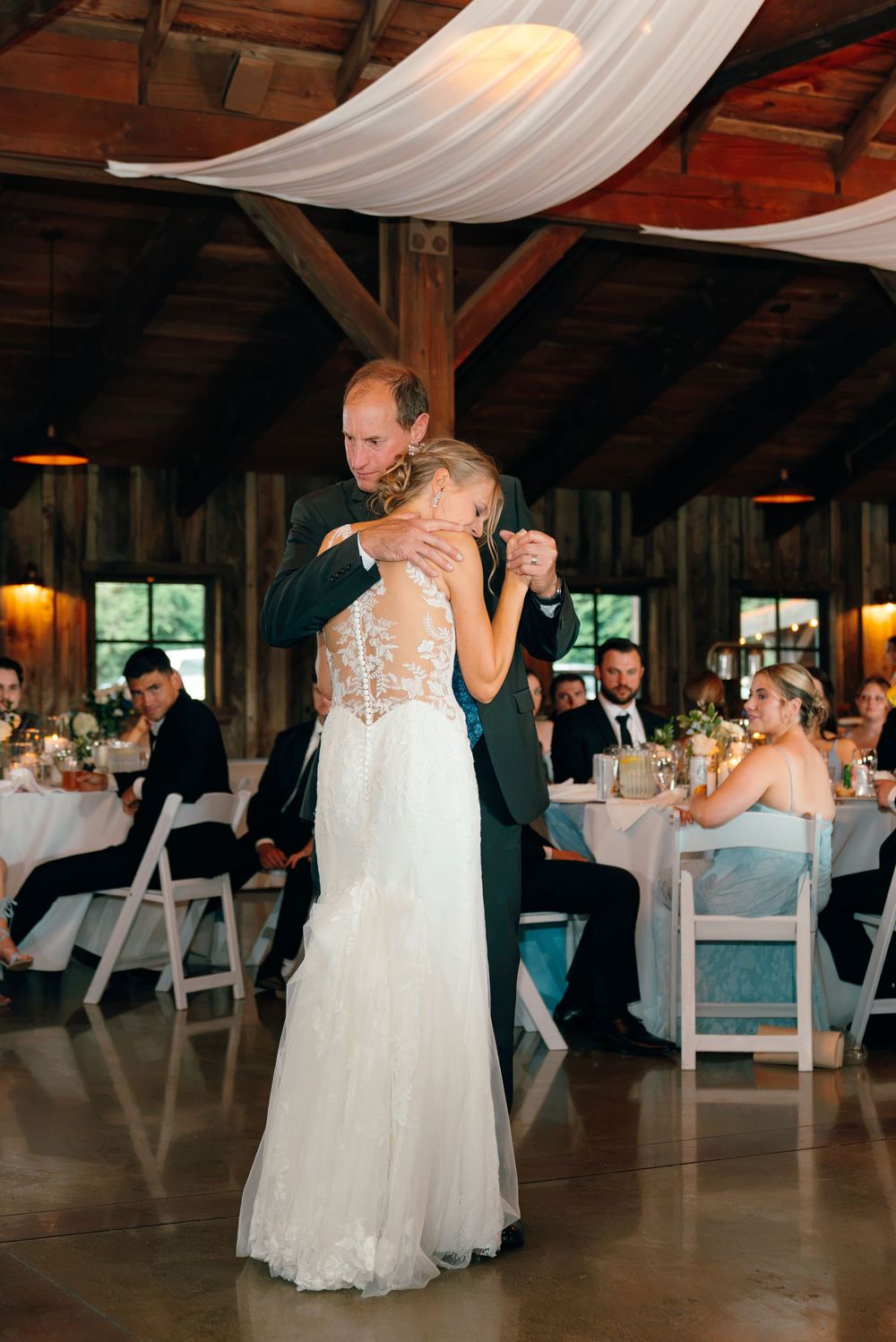 Bride and groom share their first dance in a rustic barn wedding venue with white draping and candlelit tables.