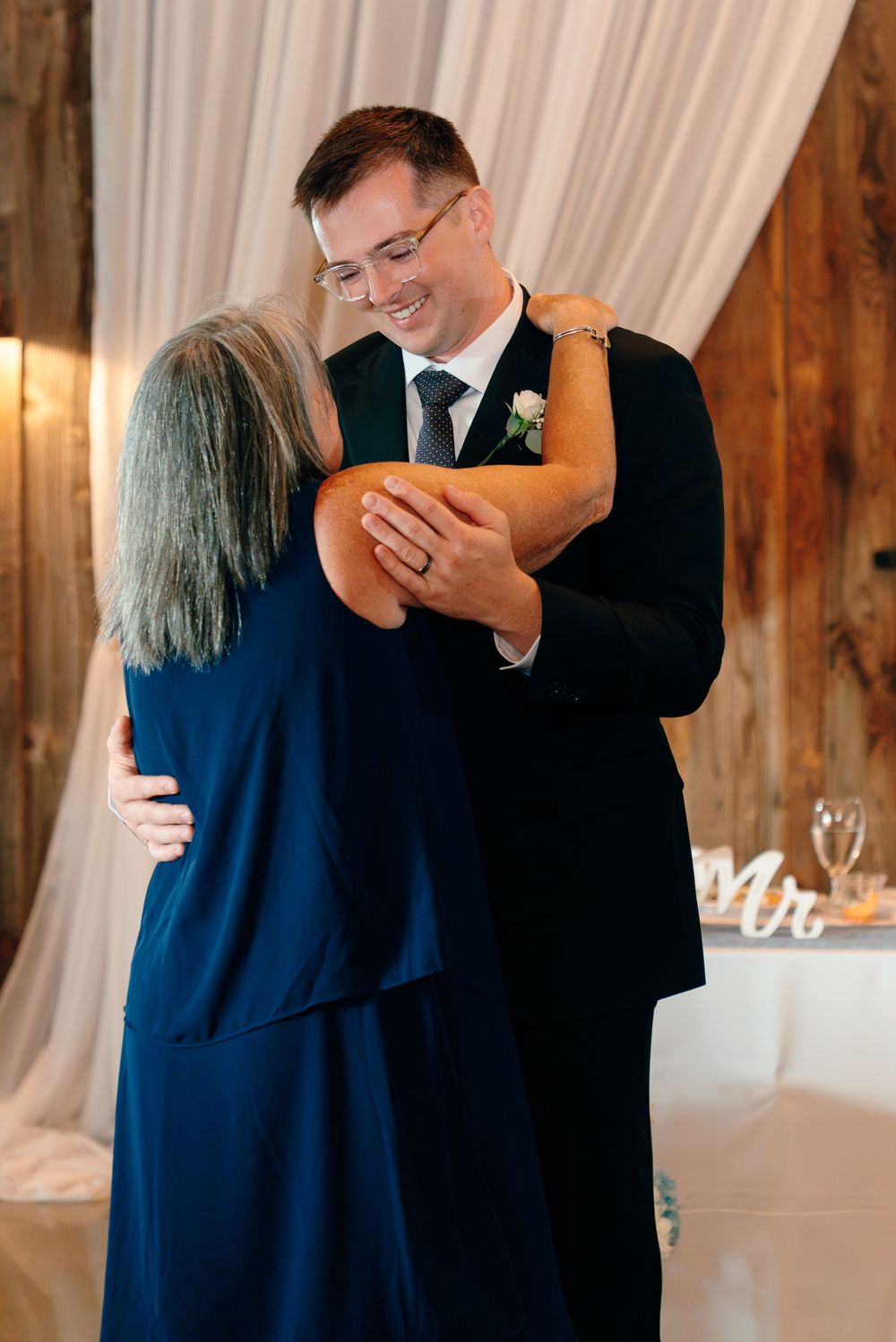 Groom in black suit shares emotional mother-son dance at rustic wedding reception, both smiling warmly.