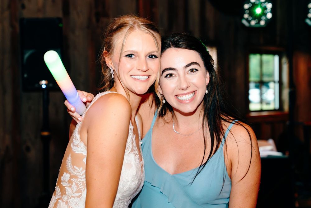 Smiling bride in lace gown poses with bridesmaid holding a glow stick at rustic wedding reception.