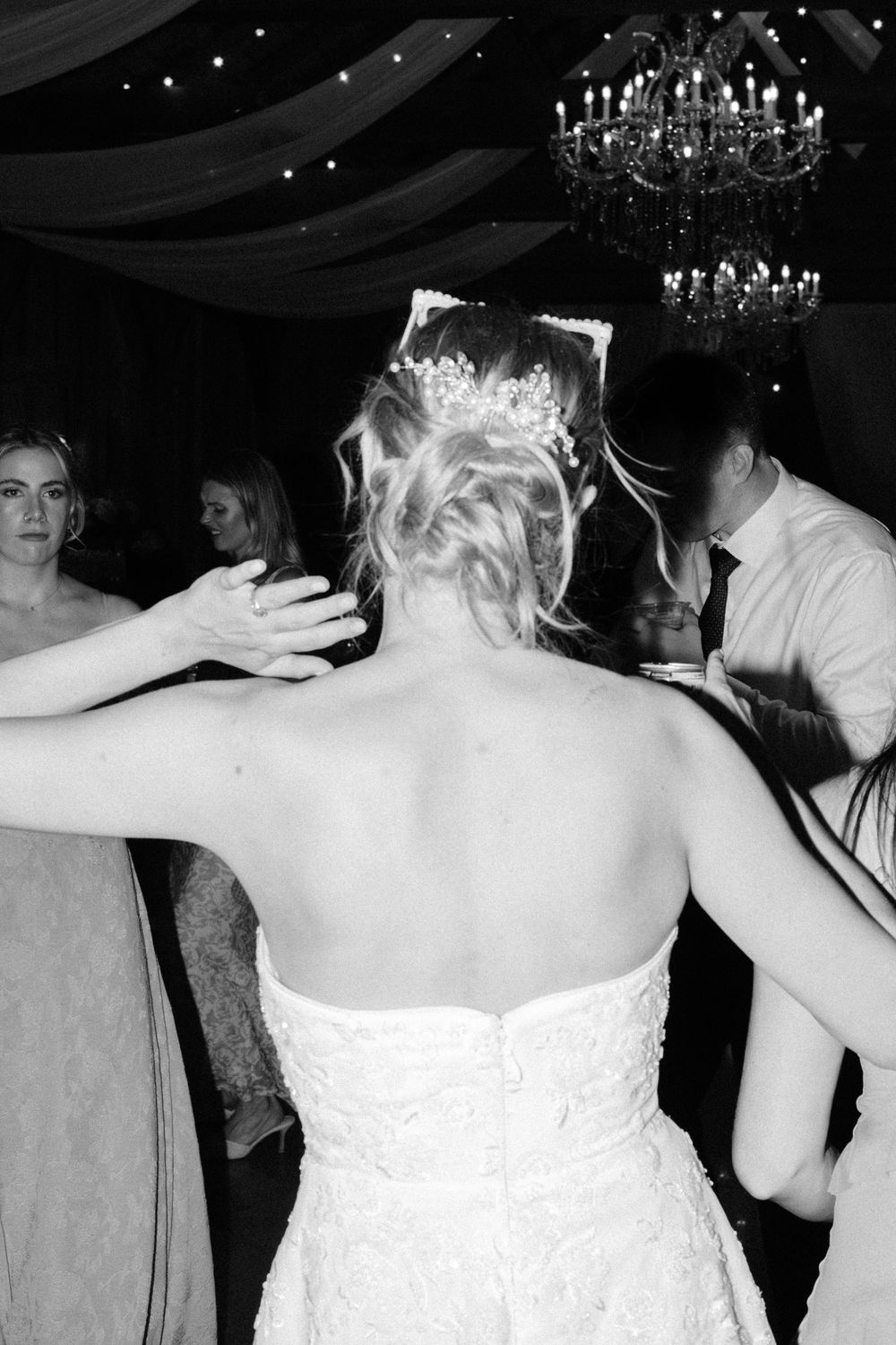 Bride with tiara updo dancing with guests under chandelier at wedding reception.
