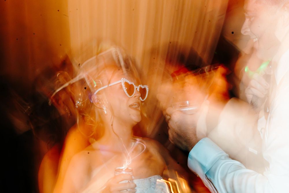 Blurred motion photo of a woman wearing heart-shaped glasses at a party, holding a drink under warm glowing lights.