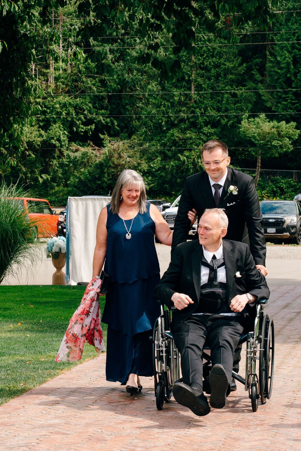 A man in a suit pushes an elderly man in a wheelchair along a brick path, accompanied by a woman in a navy dress.