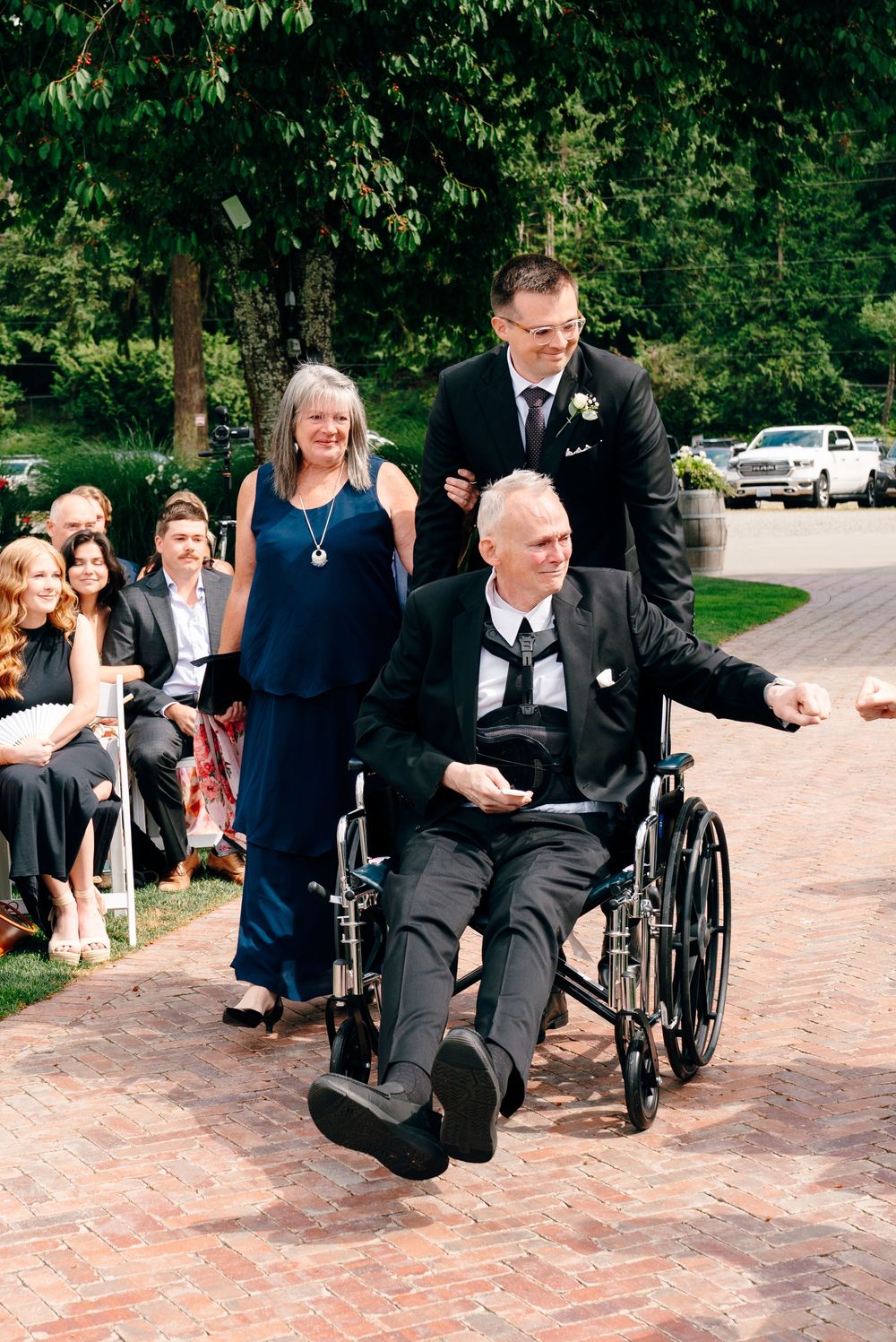 Man in wheelchair being pushed down brick aisle at outdoor wedding ceremony, accompanied by woman in navy dress.