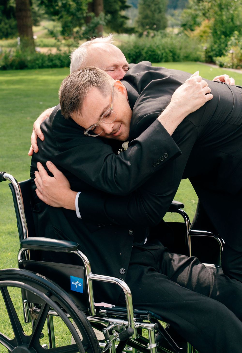 Two men in black suits share an emotional hug, one seated in a wheelchair, at an outdoor wedding ceremony.