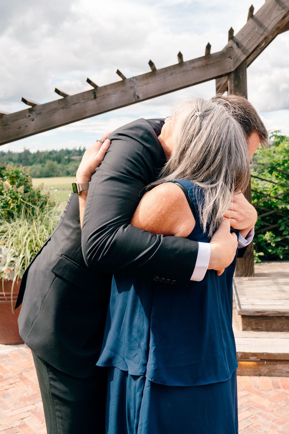 A man in a dark suit embraces a woman in a navy dress outdoors under a wooden pergola at a wedding.