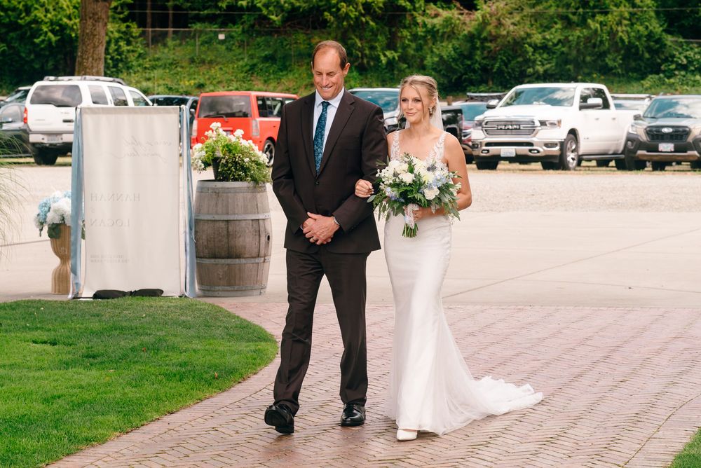 Bride in white gown holding bouquet walks with escort on brick path at outdoor wedding venue.