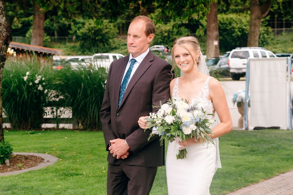 Bride in white gown holding bouquet walks with suited man outdoors at elegant garden wedding ceremony.