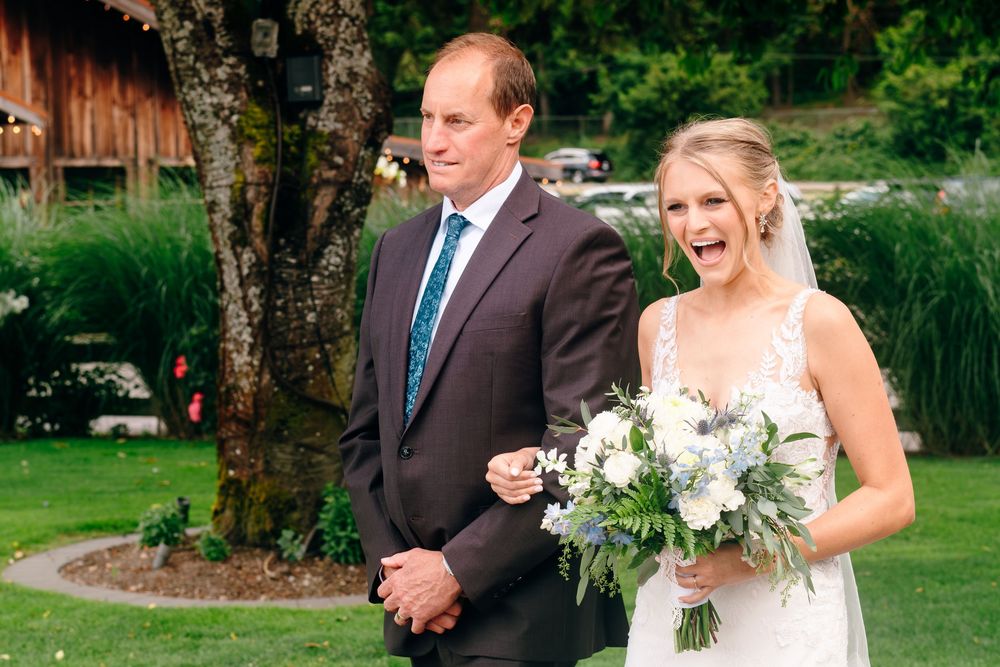 Bride in white lace gown holding white bouquet walks with man in brown suit on green lawn at outdoor wedding ceremony.