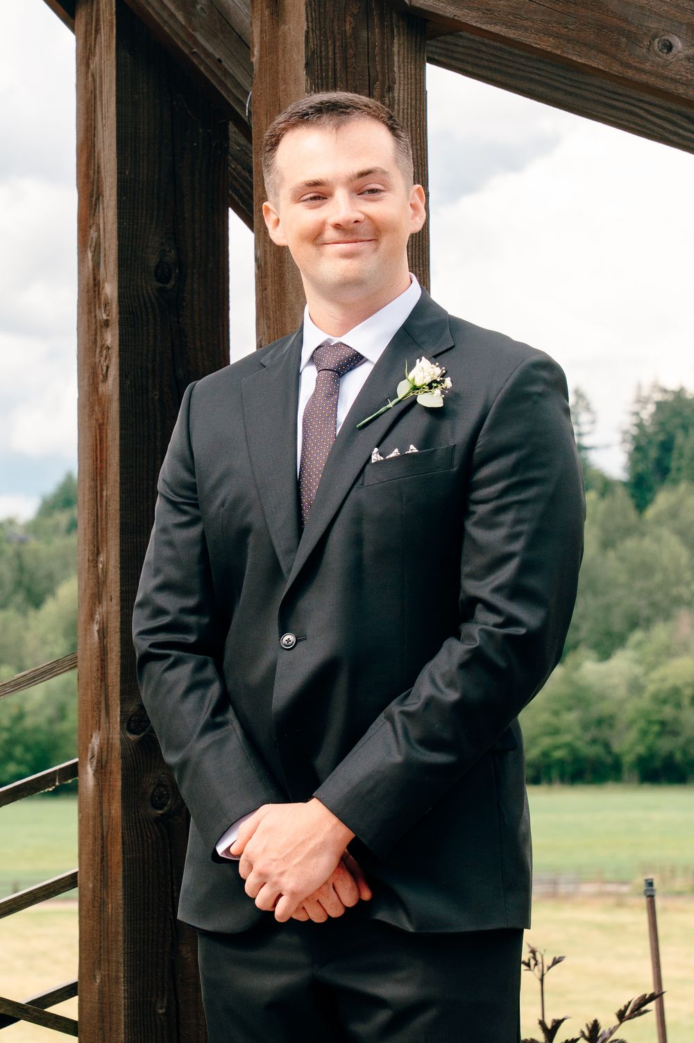 Groom in black suit with boutonniere smiling outdoors at wedding venue with green trees background.