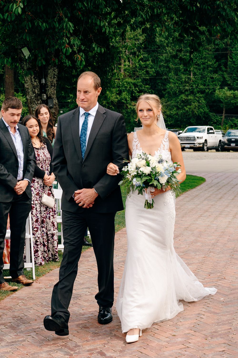 Bride in white gown holding blue and white bouquet walks down brick aisle with escort at outdoor wedding ceremony.