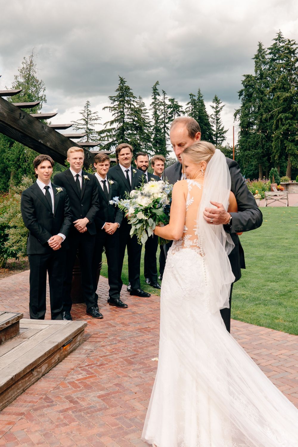 A bride in a lace gown receives a hug from a man while groomsmen in black suits look on at an outdoor wedding.