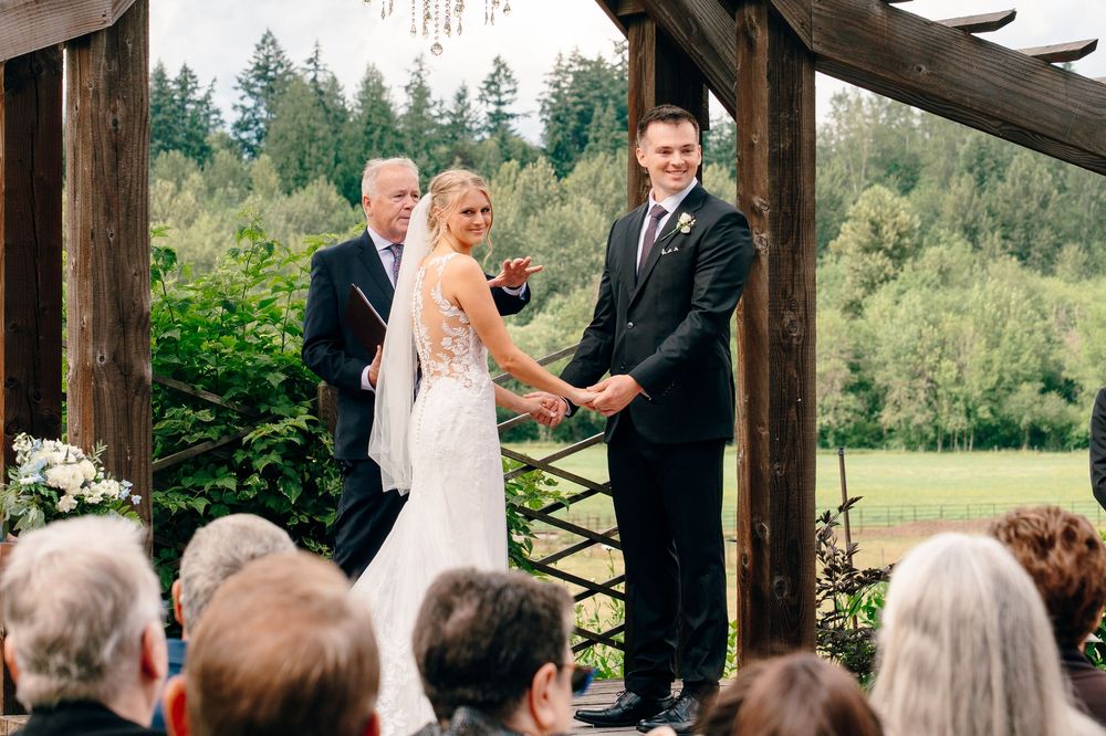 Bride and groom holding hands at outdoor wedding ceremony under wooden pergola with lush green forest backdrop.