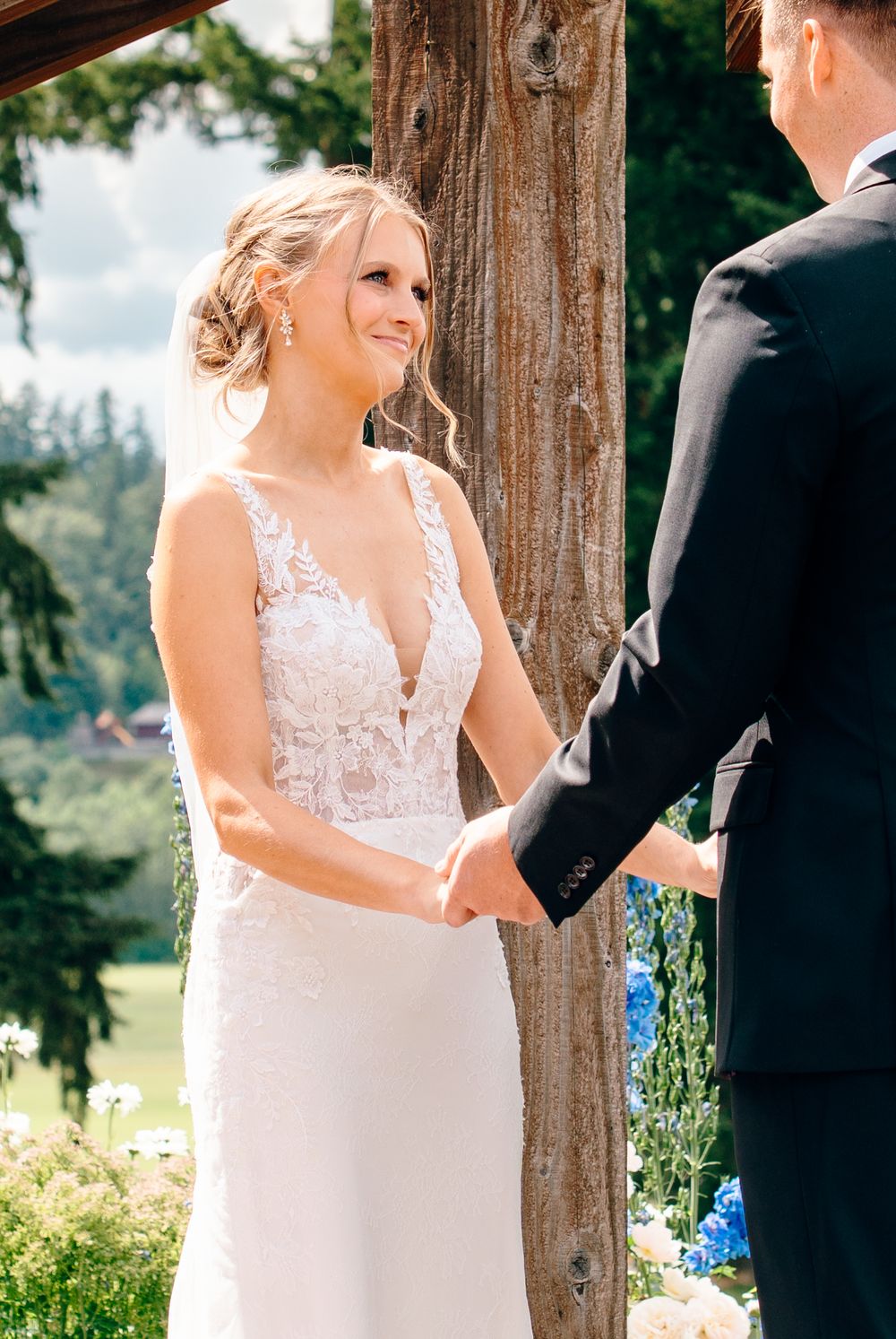 Bride in lace gown holds hands with groom during outdoor wedding ceremony, smiling joyfully beneath a tree.