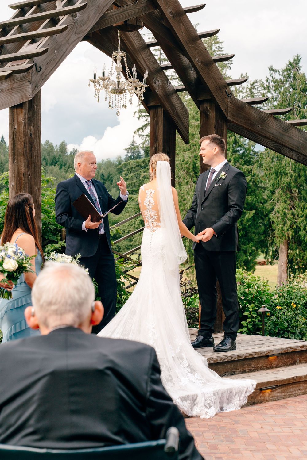 Bride and groom exchange vows under rustic wooden pergola with chandelier at outdoor garden wedding ceremony.