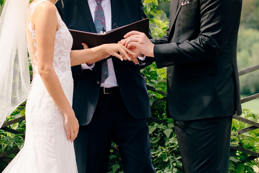 Bride and groom exchanging rings during outdoor wedding ceremony with lush green foliage in background.