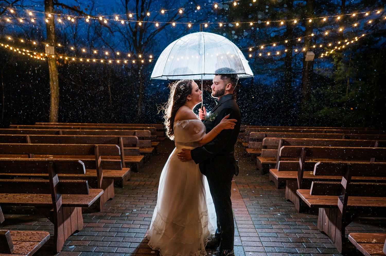 Couple sharing a romantic kiss under a clear umbrella in the rain at an outdoor wedding venue with string lights. Pavilion at Crystal Lake